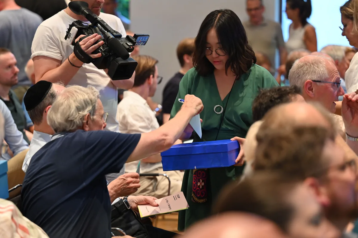 Helferinnen sammeln die Wahlzettel mit Urnen ein. Ein Wähler wirft seinen Wahlzettel in die Urne ein, die eine Helferin ihm hinstreckt.